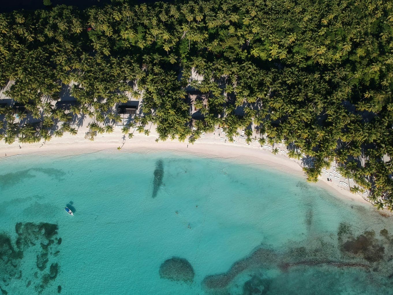 Vista aérea de una playa de arena blanca con palmeras, aguas turquesas y una pequeña embarcación